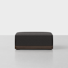 A modern, rectangular black ottoman with a dark wooden base sits on a light gray floor against a plain white background.