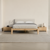 A minimalist bedroom with a light wooden bed frame, gray upholstered headboard, gray bedding, two wooden nightstands, a small lamp, a branch in a vase, and a large light gray rug on a polished floor.
