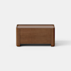 A small, rectangular wooden storage box or bench with a minimalist design, placed on a light gray surface against a plain white background.