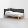 A modern daybed with a light wood frame, white mattress and pillow, and dark gray upholstered back and side panels, placed in the corner of a minimalist room with light gray flooring and white walls.