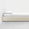 A minimalist single bed with a light beige upholstered frame, white bedding, and a pillow, placed against a white wall on a smooth, light gray floor.