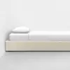 A minimalist single bed with a light beige upholstered frame, white bedding, and a pillow, placed against a white wall on a smooth, light gray floor.