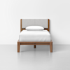 A single wooden bed with a light gray upholstered headboard, topped with white bedding and a single white pillow, set against a plain white wall and floor.