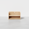 A simple, light wood storage unit with an open front, featuring one shelf in the middle and a solid back, set against a plain white background.