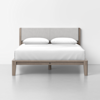 A modern bed with a light wood frame, upholstered headboard, and white bedding, including two pillows, set against a plain white background.