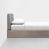 A minimalist bed with a light wood frame, grey upholstered headboard, white sheets, and a white pillow, set against a plain white wall and grey floor.