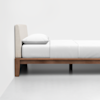 A minimalist bed with a light-colored upholstered headboard, white bedding, and a wooden frame, shown in profile against a plain white wall and floor.