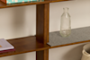 A clear, ribbed glass bottle sits on a gray felt mat on a wooden shelf. Colorful books are stacked on the upper shelf, and a woven basket is partially visible on the lower right side.