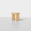 A simple wooden stool with a rectangular seat and three wide, flat legs, placed on a smooth, light gray surface against a plain white background.