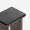 A close-up view of a modern, wooden stool with a square, dark gray cushioned seat and clean, simple lines against a white background.