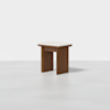A modern wooden stool with a square, flat white seat and wide, straight legs, placed on a smooth, light gray floor against a plain white background.