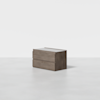 A modern, minimalist wooden cabinet with two drawers sits on a light, seamless surface against a plain white background.