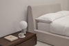 A neatly made bed with white bedding sits next to a dark wooden nightstand holding a lamp with a round white shade, a closed book, and a white cup on a saucer. The room has a minimalist design.