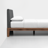 A minimalist side view of a bed with a wooden frame, gray upholstered headboard, white bedding, and a single white pillow, placed against a plain white wall and floor.
