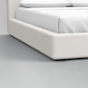 Close-up of the corner of a modern, low-profile bed with a light beige upholstered frame and white bedding, set on a light gray floor.