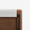 Close-up of the corner of a wooden bed frame with a light gray fabric panel attached to the top edge; the wood has a smooth finish and rounded edges.