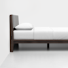 Minimalist bed with a dark wooden frame, a padded gray headboard, and white bedding, positioned against a plain white wall on a smooth, light-colored floor.