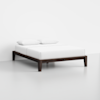 A minimalist bed with a dark wooden frame, white mattress, and two white pillows, set against a plain white wall and light-colored floor.