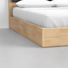 Close-up of a light wood bed frame with a white mattress and pillows, set on a smooth, light gray floor in a minimalistic room.