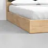 Close-up of a light wood bed frame with a white mattress and pillows, set on a smooth, light gray floor in a minimalistic room.