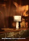 A marble table lamp with a white shade sits on a wooden shelf beside a wooden tray and a rustic vase, against a backdrop of rich wood and stone. Text reads: Stone Table Lamp, in Italian Arabescato Marble.