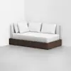 A modern daybed with a dark wooden base, white mattress, and three white pillows, positioned against two intersecting white walls in a minimalist, empty room with a light gray floor.