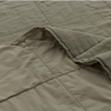 A close-up of a soft, quilted blanket in a muted khaki color, showing stitched square patterns and a slightly lifted corner revealing the texture.
