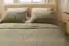 A neatly made bed with a beige upholstered headboard, two large greenish-gray pillows, and a matching quilted comforter, set in a softly lit room with cream curtains and a potted plant nearby.