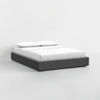 A modern, low-profile bed with a dark gray upholstered frame, white mattress, fitted sheet, and two white pillows, placed in a minimal, empty room with white walls and floor.