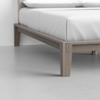 A close-up view of the corner of a wooden bed frame with tapered legs, supporting a neatly made bed with white sheets and pillows in a minimal, modern bedroom.