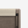 Close-up of the top corner of a wooden crib with a rounded edge and a light beige fabric covering the top rail, against a plain white background.