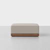 A modern, rectangular ottoman with light beige upholstery and a wooden base, placed on a smooth grey floor against a plain white background.