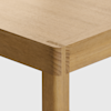 Close-up of the corner of a wooden table showing the joinery detail at the intersection of the tabletop and leg, highlighting the natural wood grain and smooth finish.