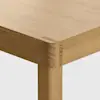 Close-up of the corner of a wooden table showing the joinery detail at the intersection of the tabletop and leg, highlighting the natural wood grain and smooth finish.