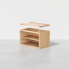 A light wood storage box with an open front and one internal shelf, shown with its flat lid lifted above it, positioned on a smooth grey floor against a plain white background.