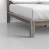 A close-up view of the corner of a wooden bed frame with light grayish-brown legs, topped with a white mattress and bedding, on a smooth light gray floor.