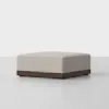 A square, beige upholstered ottoman with a dark wooden base, placed on a light gray surface against a plain white background.