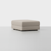 A large, square, light beige ottoman with clean lines and a minimalist design, placed on a light gray floor against a plain white background.