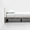A modern, minimalist bed with a light-colored upholstered headboard, white bedding, and a single white pillow, set against a plain white wall and floor.