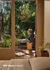 A person sits on a wooden bench on a sunlit outdoor deck, surrounded by green plants and trees. The scene is viewed through an open doorway from inside a cozy room. Text reads: Pillar Bench, in Natural.