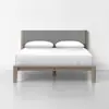A modern bed with a light wooden frame, a gray upholstered headboard, two white pillows, and a white bedspread, set against a plain white wall and floor.