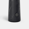 A close-up of the base of a cylindrical object made of dark marble with subtle white veining, featuring a small round metallic button or knob on its side, set against a plain light background.