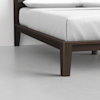 Close-up of the corner of a dark wooden bed frame with neatly made white bedding, positioned on a smooth light gray floor against a white wall.