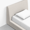 A close-up of a modern bed with a light beige upholstered headboard and frame, white bed sheets, and a single white pillow, placed on a gray floor against a white wall.
