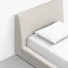 A close-up of a modern bed with a light beige upholstered headboard and frame, white bed sheets, and a single white pillow, placed on a gray floor against a white wall.