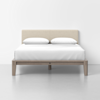 A modern bed with a light beige upholstered headboard, two white pillows, and a neatly made white bedspread, set against a plain white wall and light gray floor.