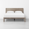 A wooden bed frame with a rectangular headboard, fitted with a white mattress and two white pillows, stands on a plain, light-colored floor against a white wall.