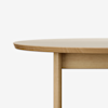 Close-up of the edge and leg of a round, light wood table against a plain white background. The image highlights the smooth finish and simple, minimalist design of the table.