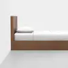 A minimalist wooden bed frame with a tall rectangular headboard, white pillow, and neatly made white bedding, positioned against a plain white wall and light gray floor.