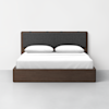 A modern wooden bed with a dark frame, a gray upholstered headboard, white bedding, and two white pillows, set against a plain white wall and light gray floor.